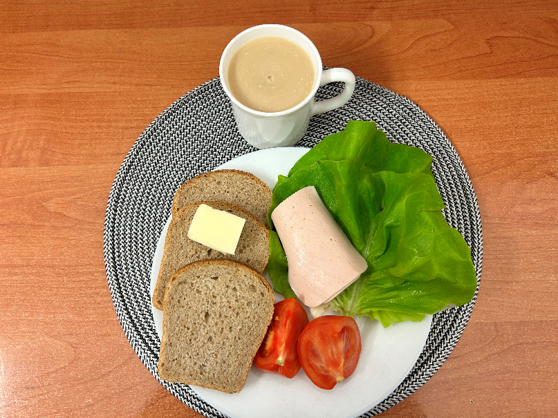 Na zdjęciu znajduje się: Kawa z mlekiem, Chleb Graham, Masło extra 82%, Szynka Drobiowa, parzona, z dodatkiem białka wieprzowego, ​​​​​​​Sałata zielona, Pomidor